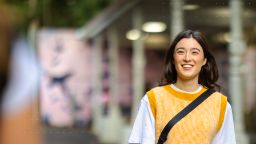 Smiling student walking on campus wearing a yellow knit vest and white t-shirt, with a blurred background of university buildings.