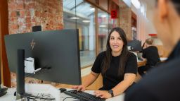 A Swinburne staff member smiles while assisting a visitor at the Future Student HQ reception desk on Hawthorn campus.