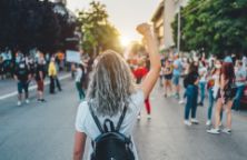The view from behind a young woman wearing a backpack with a raised fist protesting in the street.