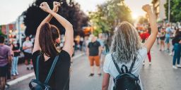 The view from behind two young women wearing backpacks with their fists raised protesting in the street.