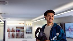A smiling young man with curly dark hair and a moustache stands in a modern medical education lab, holding a VR headset. He wears a teal and navy Adidas jacket over a cream t-shirt. Behind him are digital anatomy tables, anatomical display cabinets, a large screen showing a body scan, and a human skeleton model. The room is lit with recessed ceiling lights.