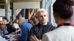 A photo of a crowd at Swinburne's Study Expo inside a modern venue on Hawthorn campus, with concrete pillars and high ceilings. In the foreground, a young man with bleached blond hair, wearing a dark jacket, looks toward the camera as he converses with a woman whose back is to the viewer. Behind them, a diverse group of attendees mingles and examines printed materials on round tables. Colourful artwork is visible on the wall in the background. 