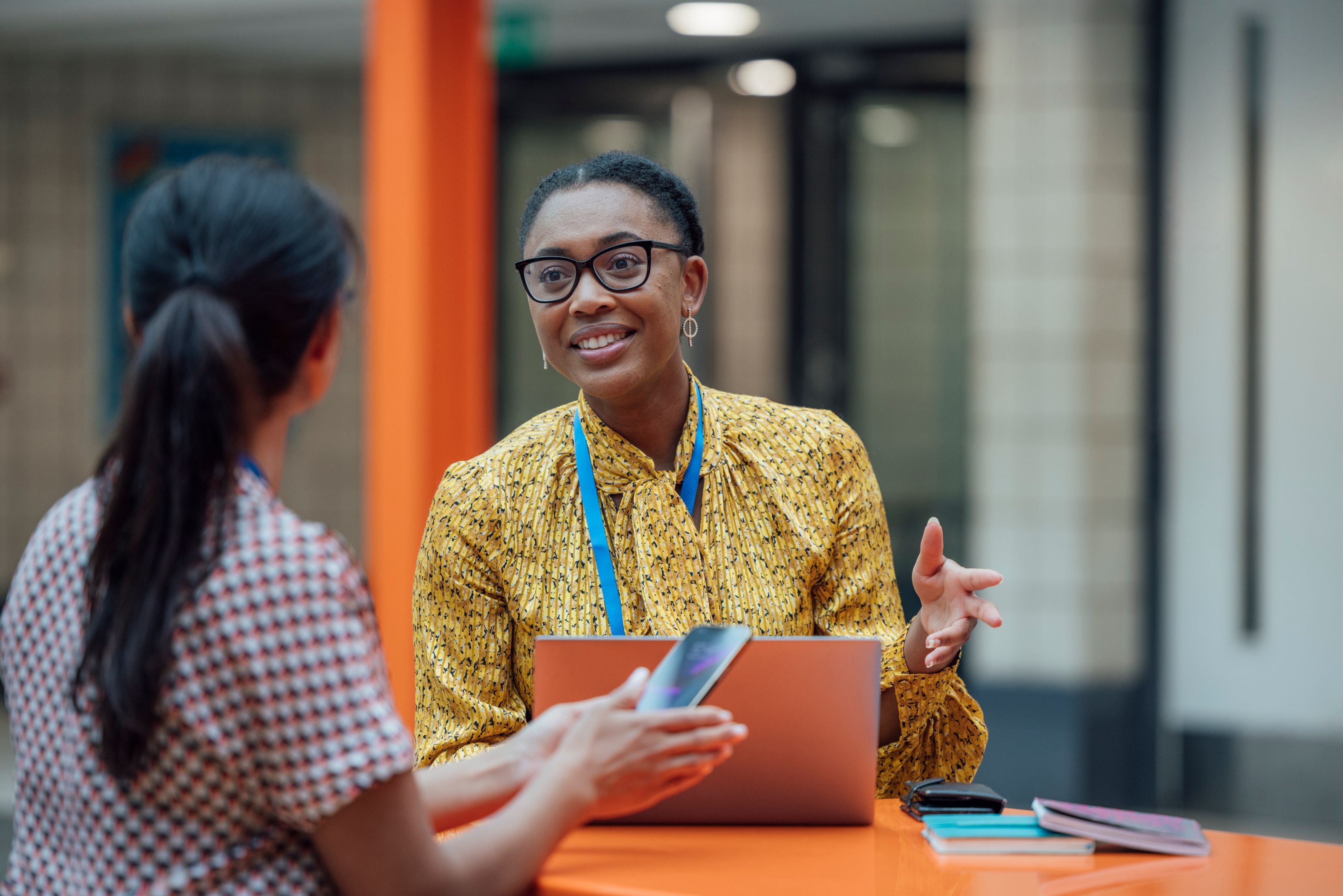 A secondary school teacher sits across from a student, smiling and gesturing as they talk. The teacher wears glasses and a yellow patterned blouse with a blue lanyard and holds a laptop. They are seated at an orange table in a bright, modern school building.