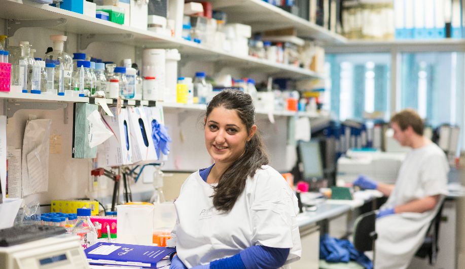 A female student on professional placement, wearing healthcare attire, rubber gloves and in front of shelves of medicine. 