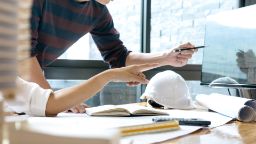 Two professional engineers sit at desk with hands pointing to computer screen. A hard hat and ruler are on the desk.