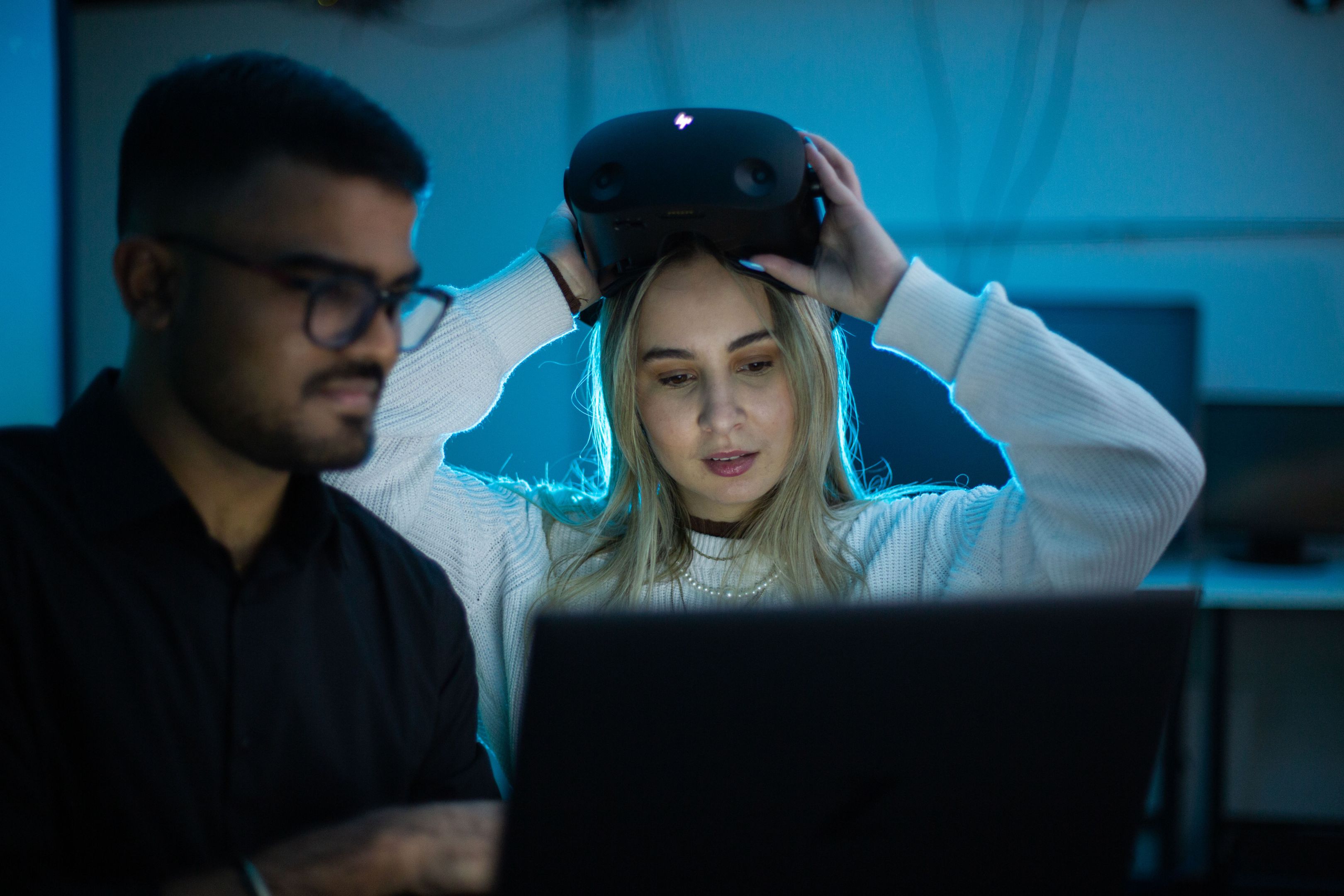 Two Swinburne computer science students collaborate in a darkened lab, one lifting a VR headset while reviewing work on a laptop..