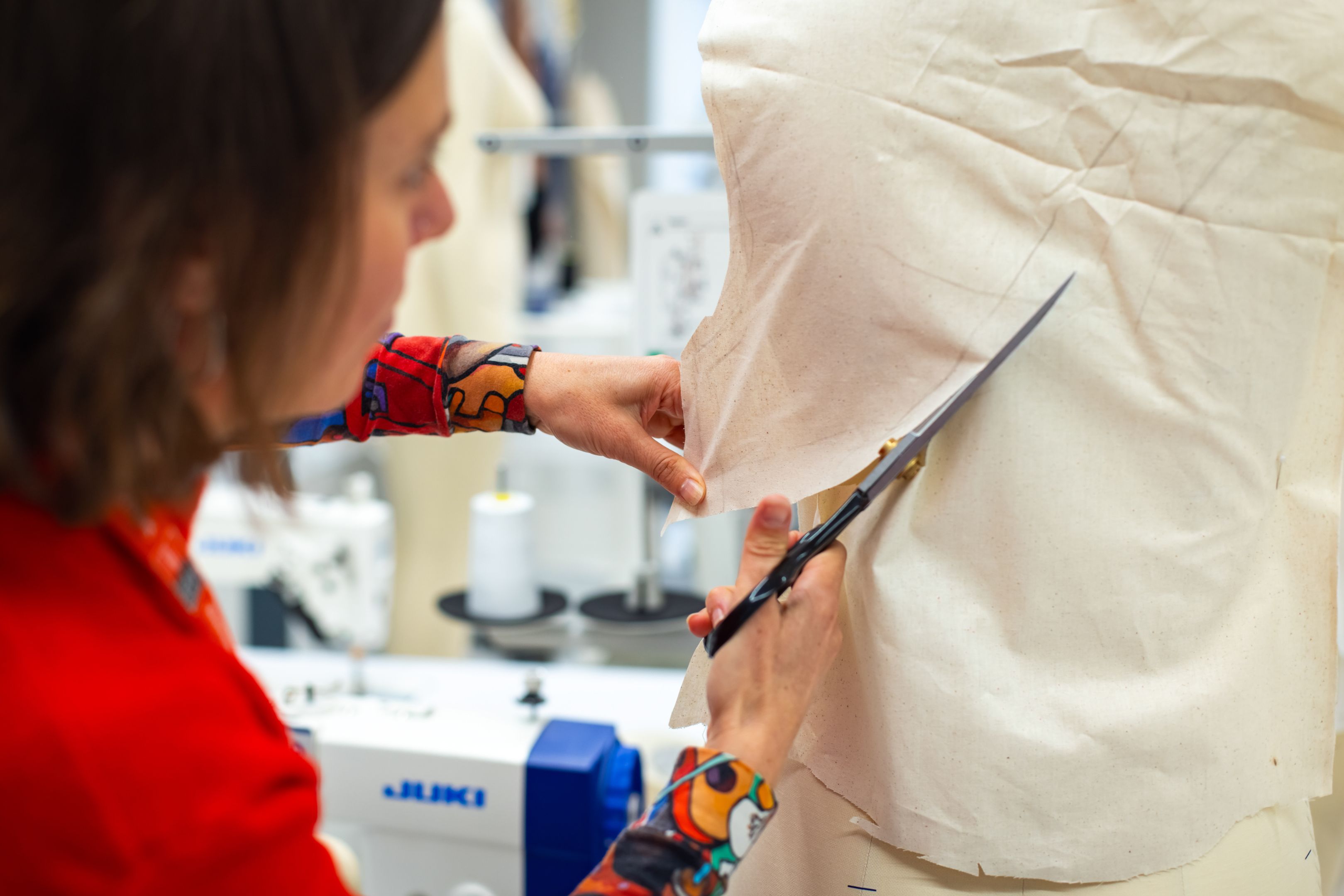 A fashion design student cutting cream-coloured muslin fabric draped on a dress form, using large tailor's scissors. She wears a vibrant red top with a colourful printed sleeve. In the background, a Juki industrial sewing machine and spools of thread are visible in what appears to be a professional atelier or fashion studio.