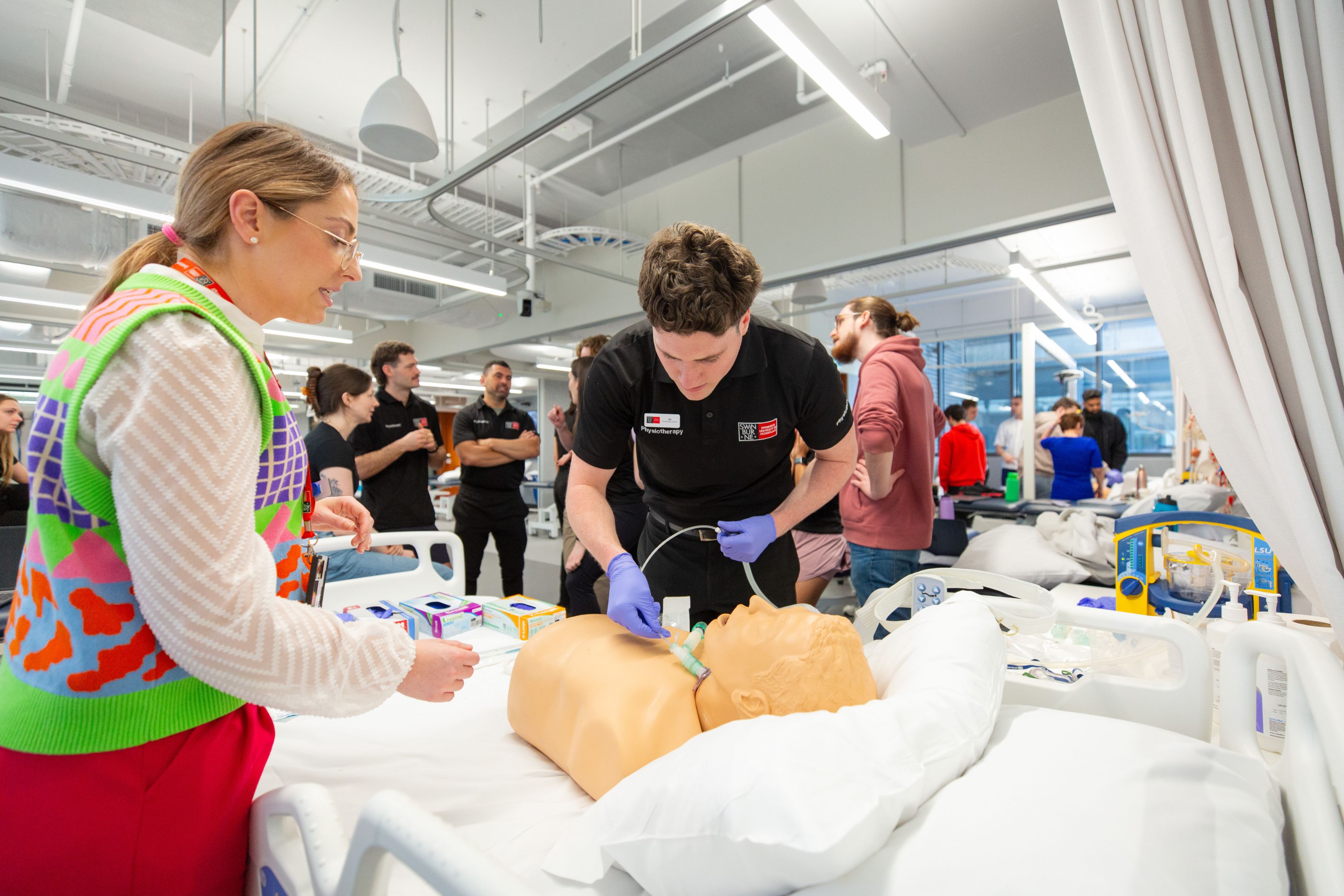 A physiotherapy student wearing blue gloves practises airway management on a medical training manikin in a clinical simulation lab. A smiling instructor in a colourful knit vest looks on. In the background, other students work at additional simulation stations equipped with medical equipment and defibrillators. The modern facility features white curtain dividers and bright overhead lighting.