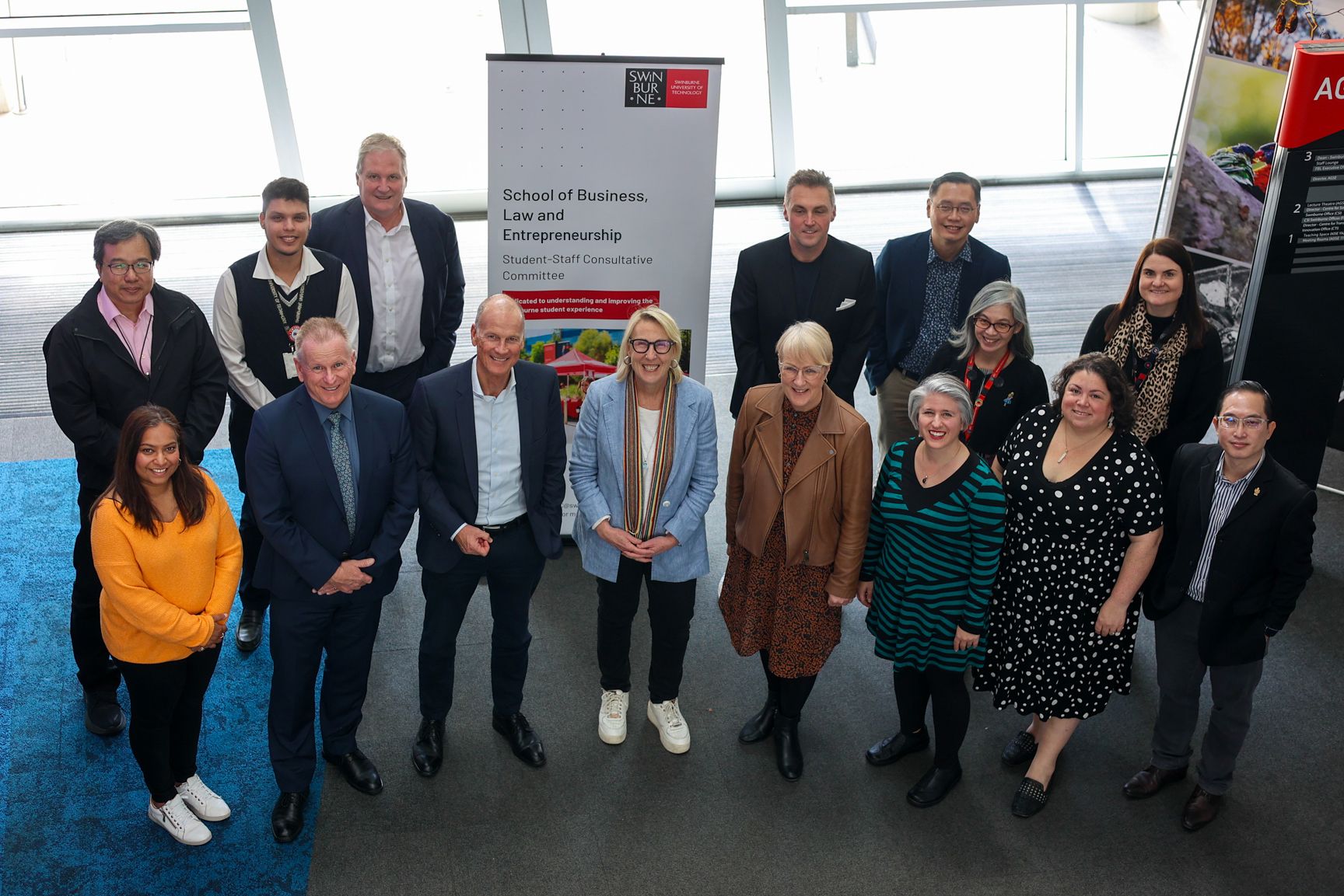 15 people standing on a lower level from the camera in a foyer, smiling at the camera infront of a banner. 