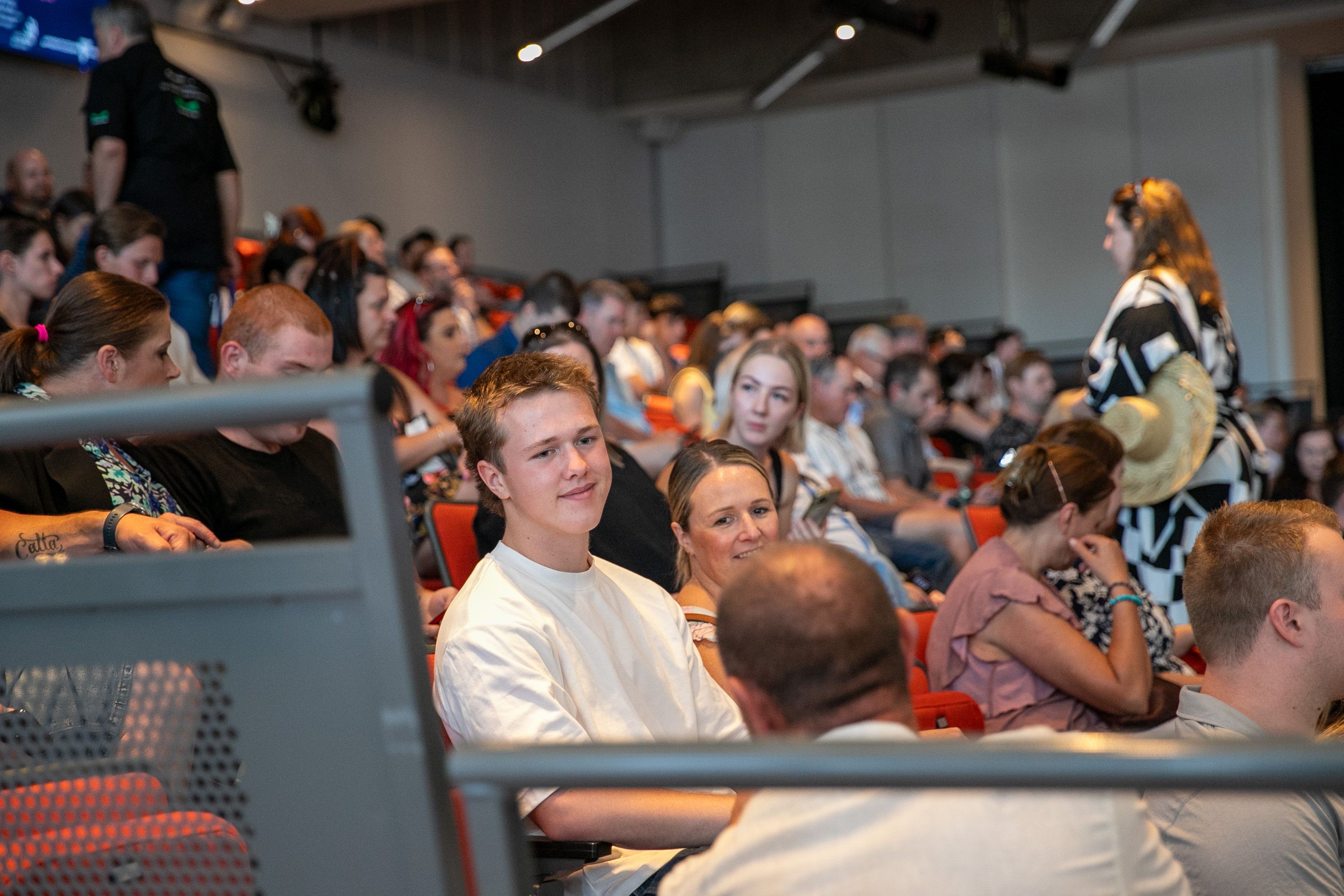 Man is sitting in an crowded auditorium smiling and looking at a person sitting in front of them, having a conversation. 