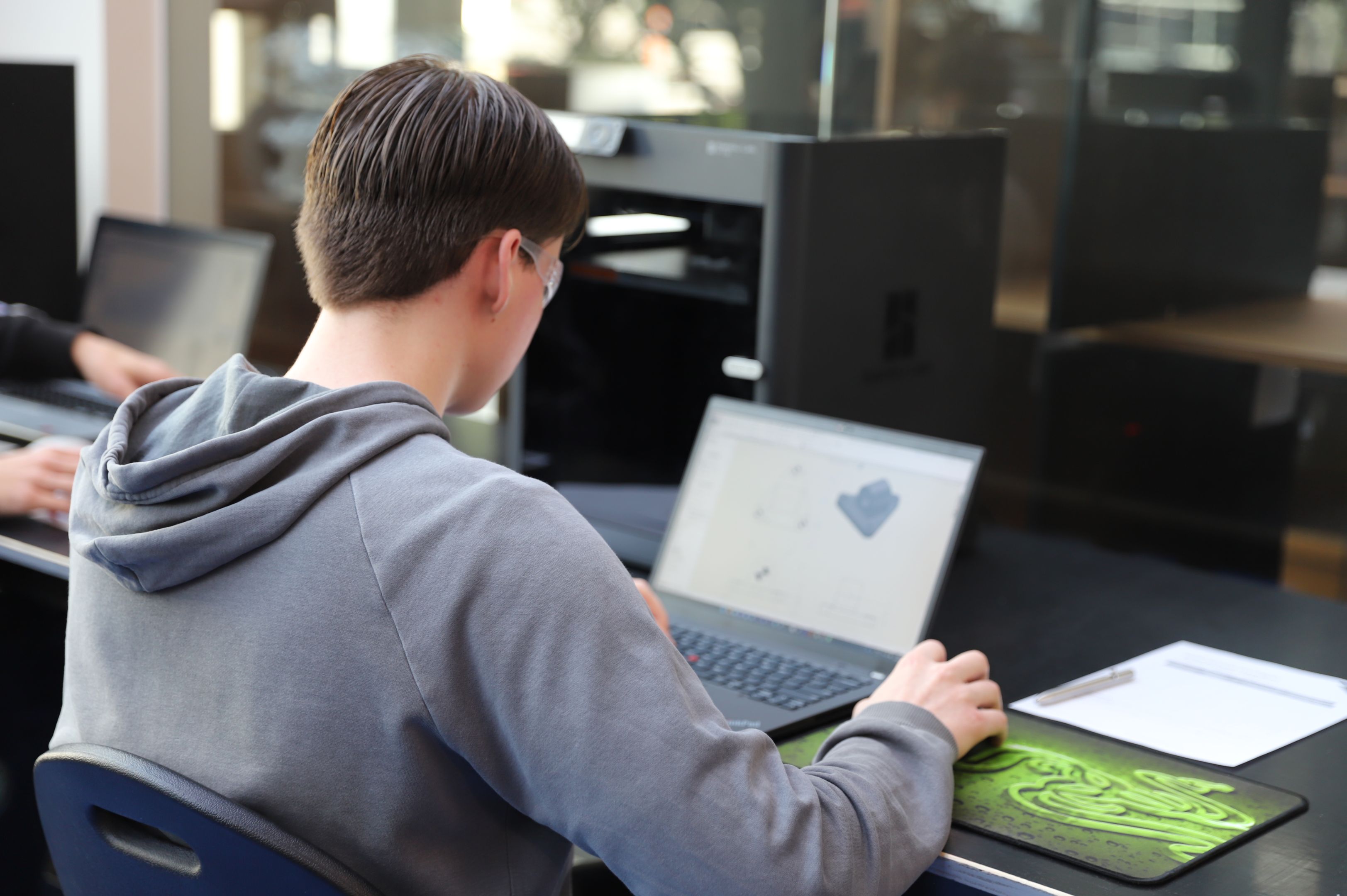 Young male student sitting at a laptop at a desk, their body faced away from the camera, wearing goggles and with their hand on a mouse. 