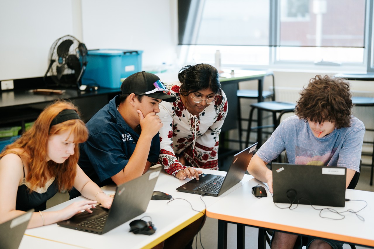 Three students and a teacher gathered at desks on laptops in a bright classroom.