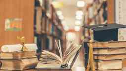 Mortarboard graduation cap and testamur sitting amongst a stack of books.