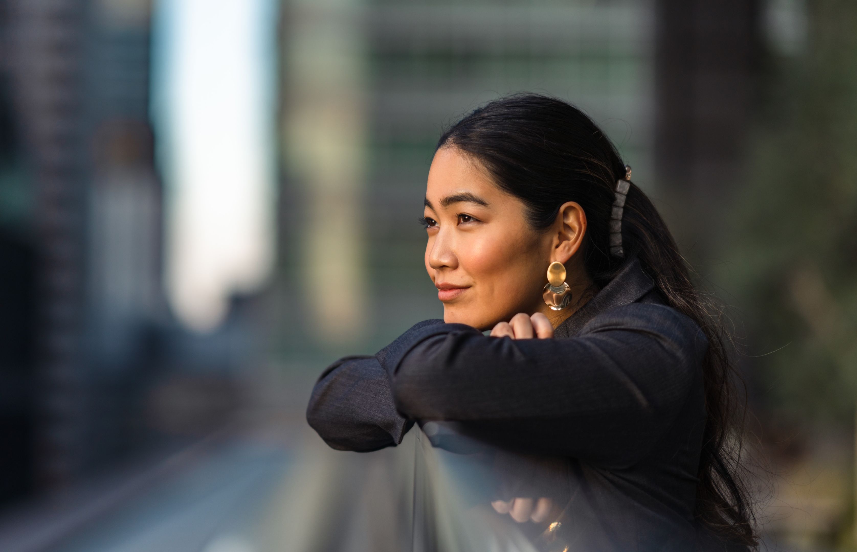 A brunette woman leans on a guard rail in an urban area and stares off into the distance.