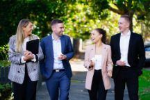 Colour image of four people two men and two women, walking down a park carrying a laptop and coffee, smiling and chatting.