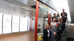 A group of Swinburne staff all standing together on a staircase with glass railings to the right of an empty room with wooden floors and sticky notes stuck to the windows. 