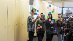 A group of EIX staff stand in front of a glass wall in an office with sticky notes on it conducting a group brainstorming and design thinking activity