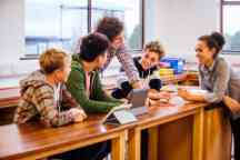 Students with their teacher sat at a table in the science lab using a digital tablet. They are caually dressed.