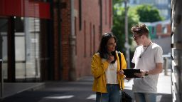 2 students standing together looking at tablet and talking, on McLeod Lane, next to the AMDC and FS buildings.