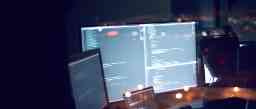 Computer monitors displaying lines of code in a dimly lit workspace.