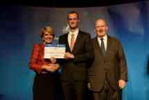 Three people stand side by side — a woman in a red jacket is on the left, shaking hands with a tall young man wearing a suit who is in the middle, holding a certificate. To their right is an older man wearing a suit. All three are smiling for the camera in front of a dark blue background. 