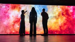 3 people stand in front of a large digital screen showing swirling images of space in red, pink and yellow. 