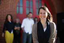 Caucasian female with brown hair smiling at the camera with a group of people semi in focus behind her