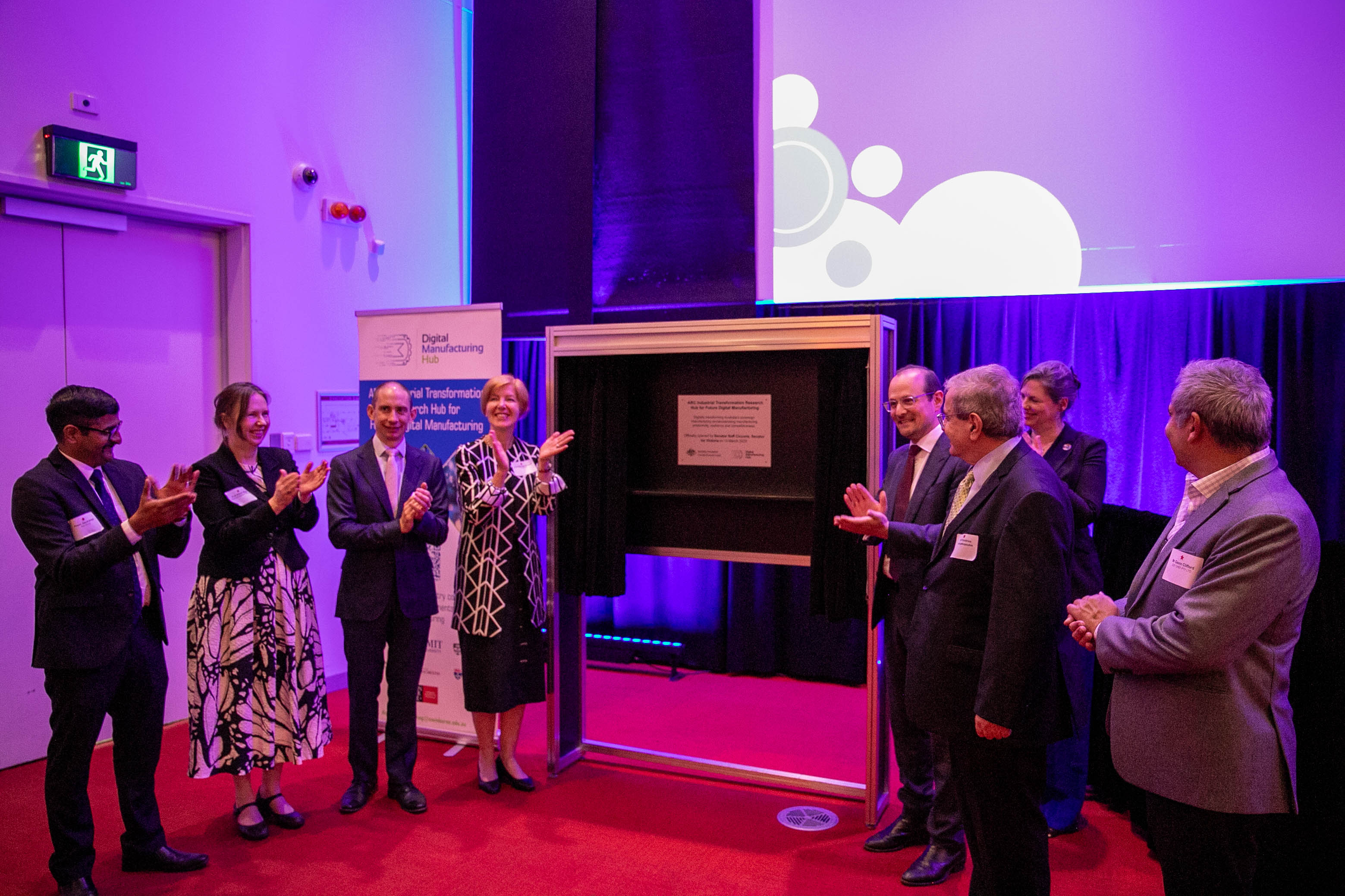 Eight guests stand beside a newly unveiled plaque at the Swinburne-led Digital Manufacturing Hub, clapping.