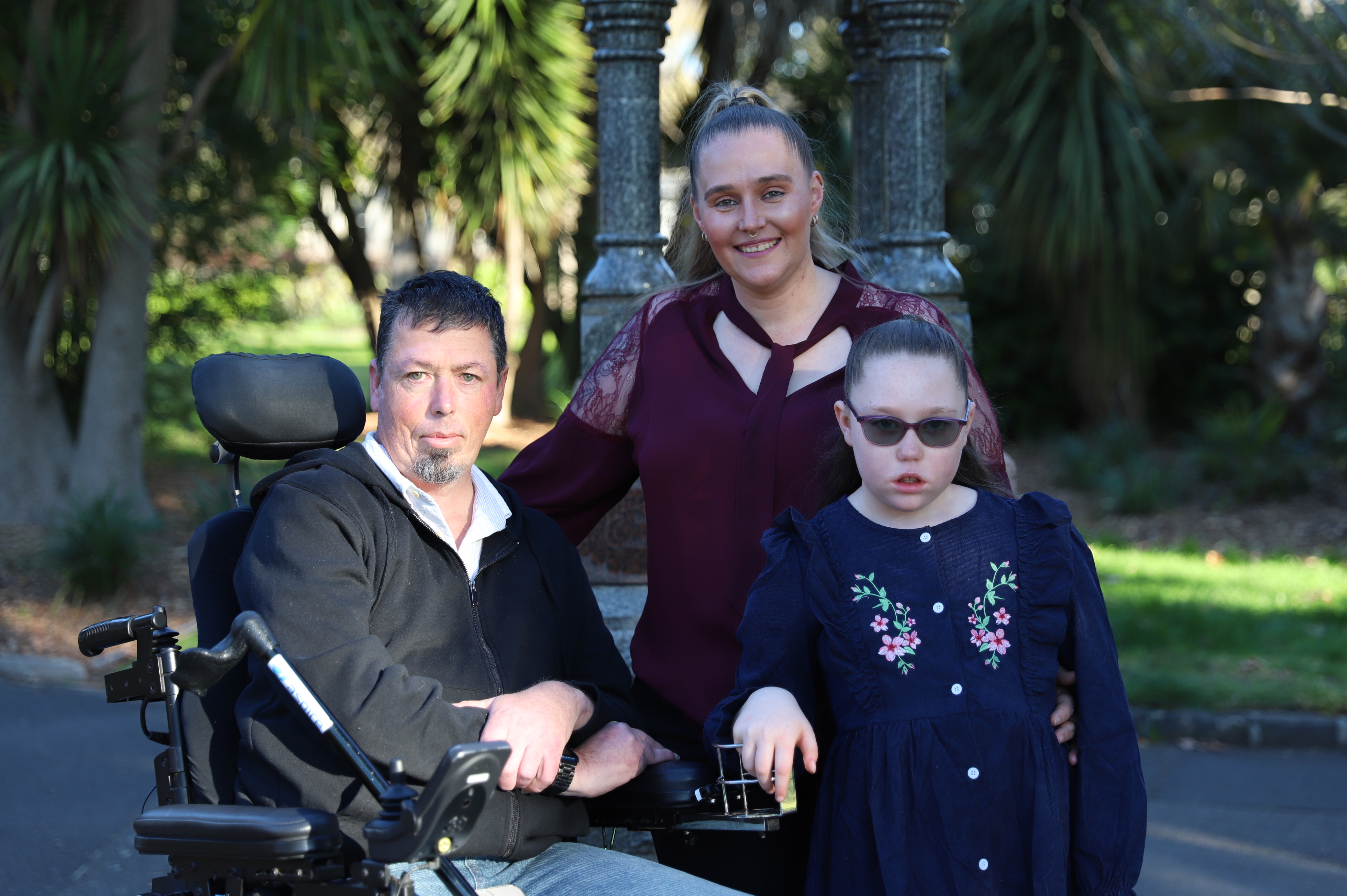 Mark, Dimity, and Zaharrah Priestley smile at the camera, with parkland behind them.