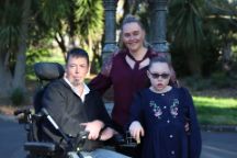 Mark, Dimity, and Zaharrah Priestley smile at the camera, with parkland behind them.