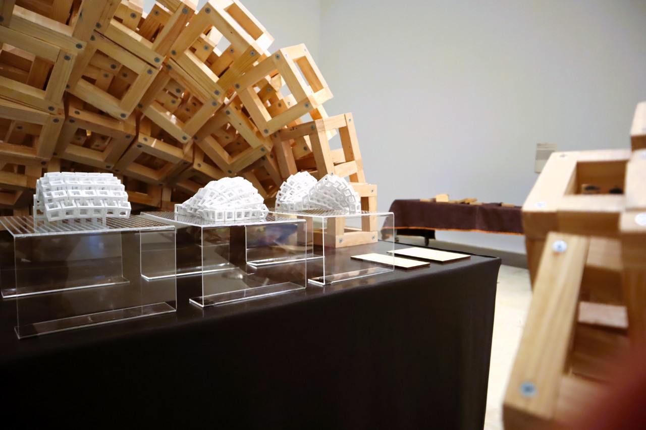 Three small plastic prototypes, made from interlocking cubes, form wavy shapes. A much larger wooden version of the same prototype sits in the background