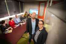 A man and a woman smiling at the camera with a group of works chatting at a table behind them