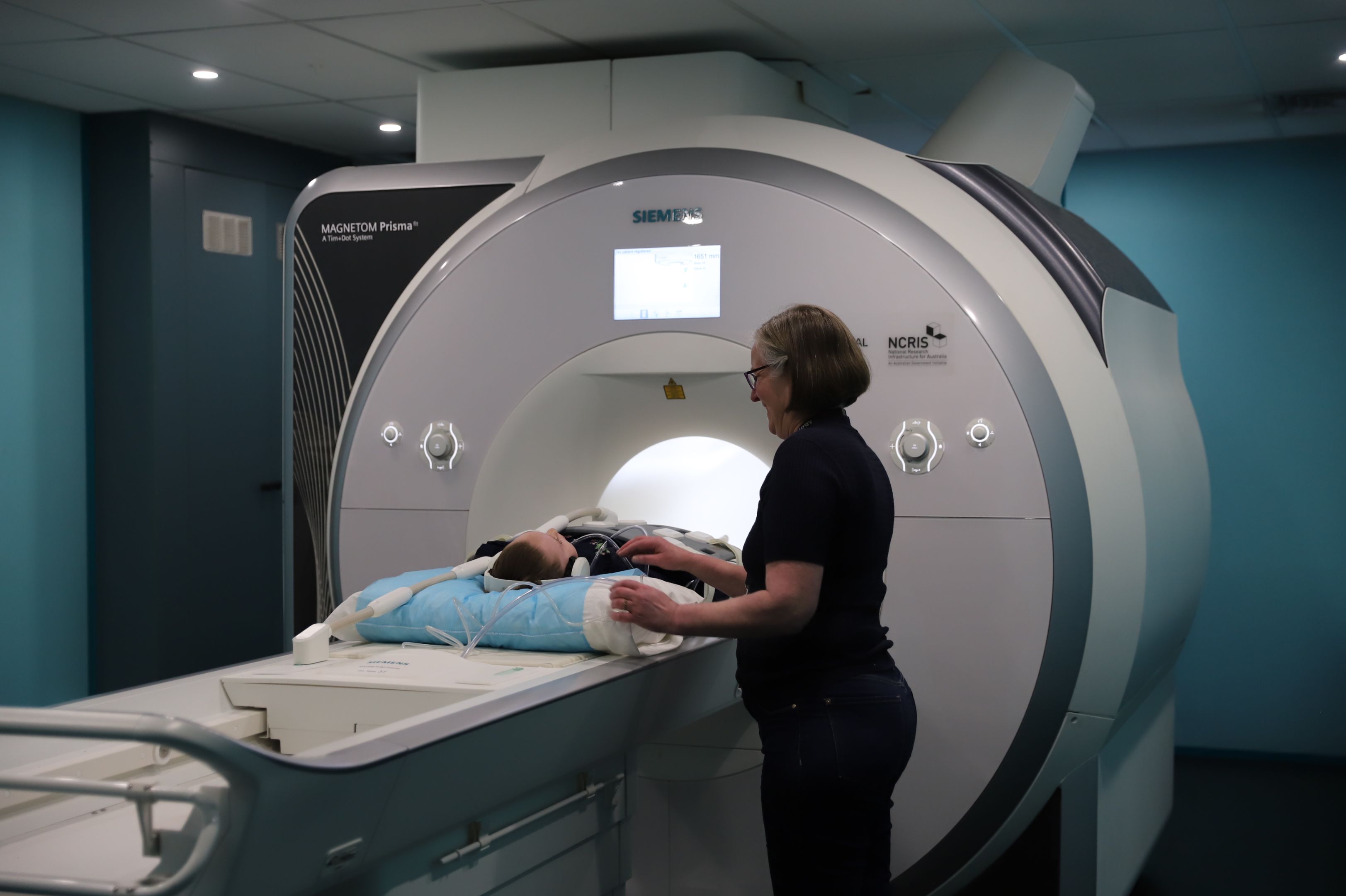 A young girl lies half-way inside an MRI machine, with a technician standing beside her.