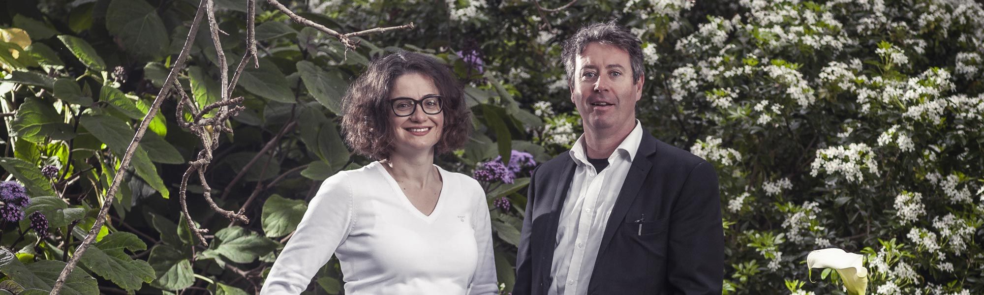 Professor Niki Frantzeskaki wearing a white top on the left and Dr. Stephen Glackin wearing a dark blazer on the right, both pictured in front of a luscious green garden.