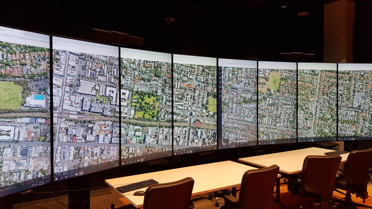 Inside a large room two long tables with four office chairs behind them are set up in front of a wide curving wall of 8 tall screens, all displaying a single image that is an aerial view of houses and streets.