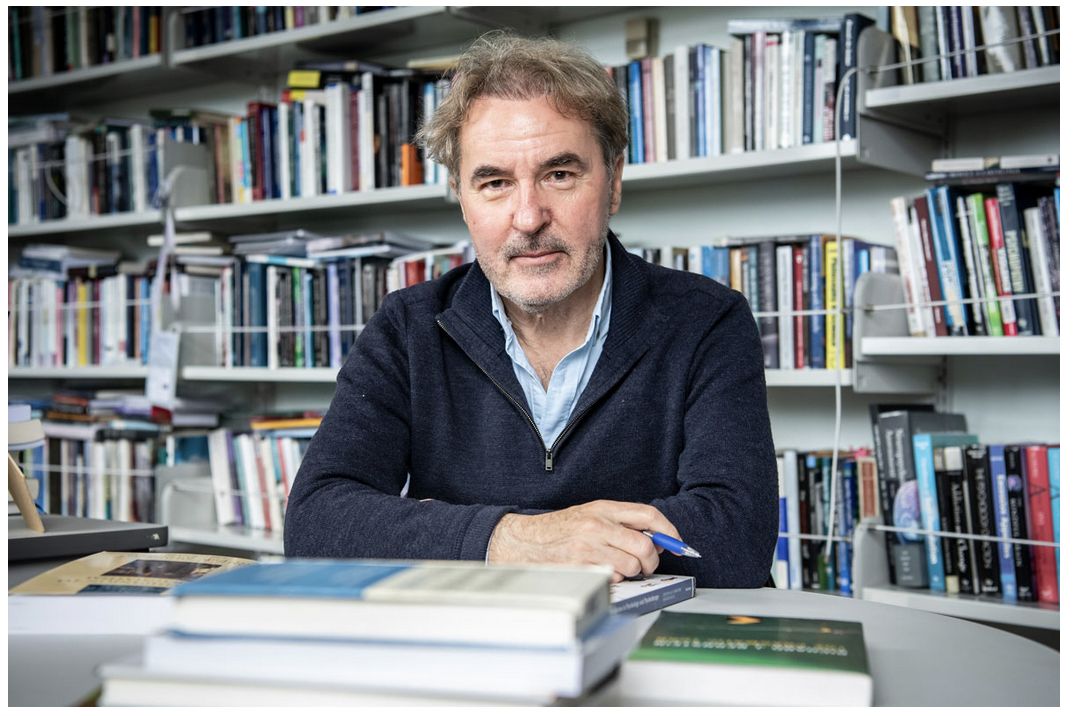 Headshot of man in navy quarter-zip jumper and blue shirt holding a pen, sitting at a desk in front of bookshelves