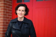 Woman with short brown hair in a black trench coat standing in front of red door.