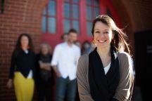 Woman with brown hair and cardigan with black top smiling at the camera, group of people  standing behind her blurred out.