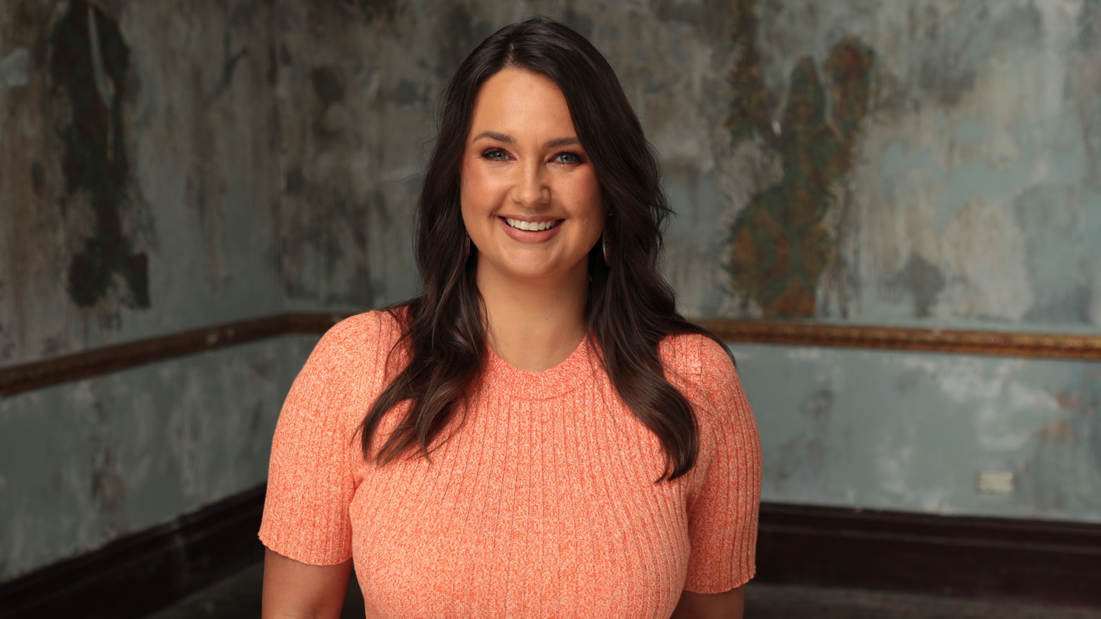 Rae Johnstone stands smiling warmly into the camera. She has long dark hair and is wearing a bright peach-coloured dress