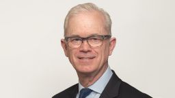 Professional looking man in a suit wearing glasses, standing slightly side on, facing the camera and smiling. 