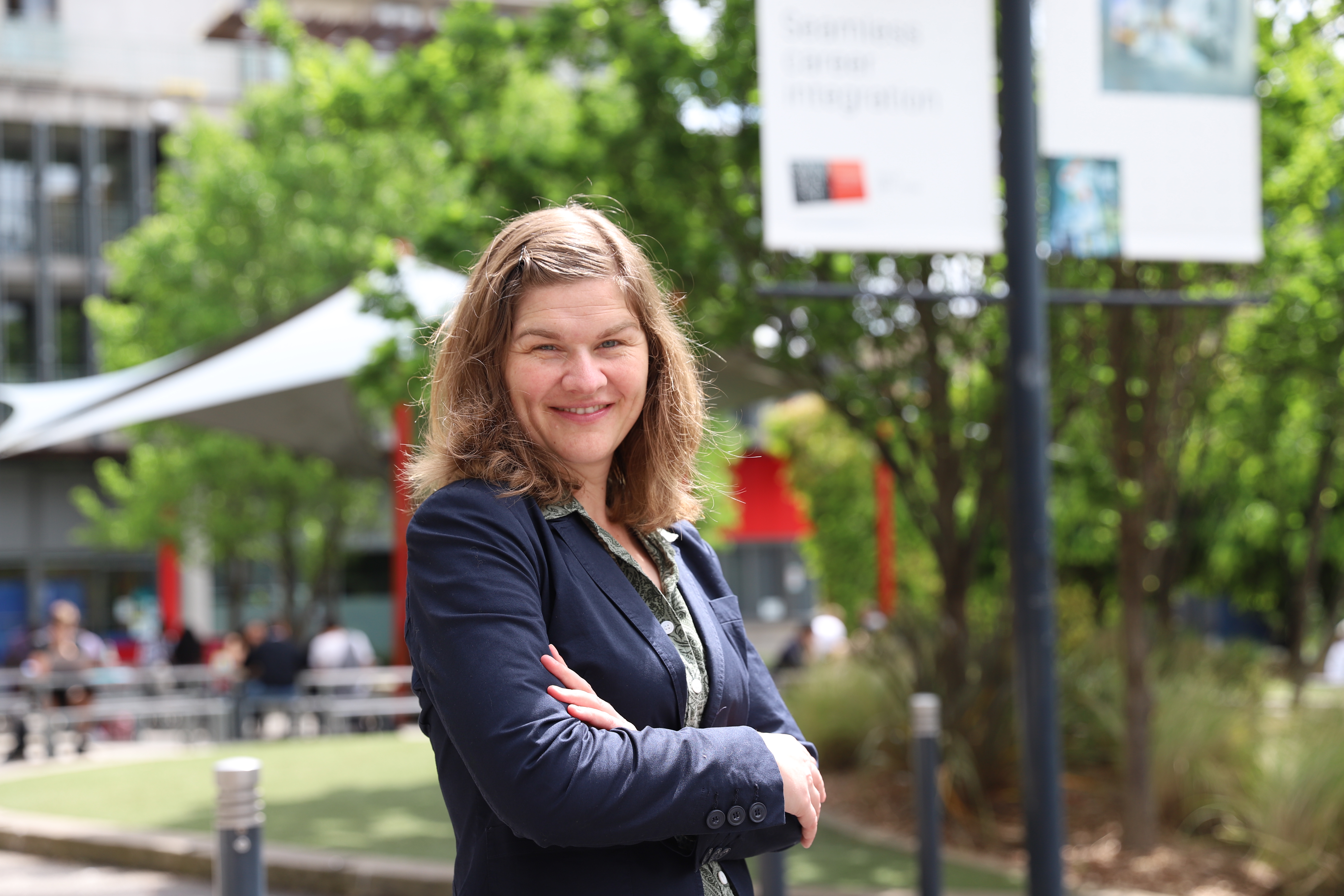 Swinburne’s director of student engagement Nadine Zacharias stands with her arms crossed, smiling into the camera. She wears her shoulder-length hair down and wears a blazer. She stands outside in the sunshine on Swinburne’s Hawthorn campus, with students and staff enjoying the good weather in the background.vvvvvvvvvvvvvvvv