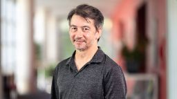 Man in a grey shirt facing slightly side on, looking at the camera and smiling.