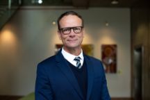 A male with short hair and glasses wearing a blue suit, white shirt and tie.