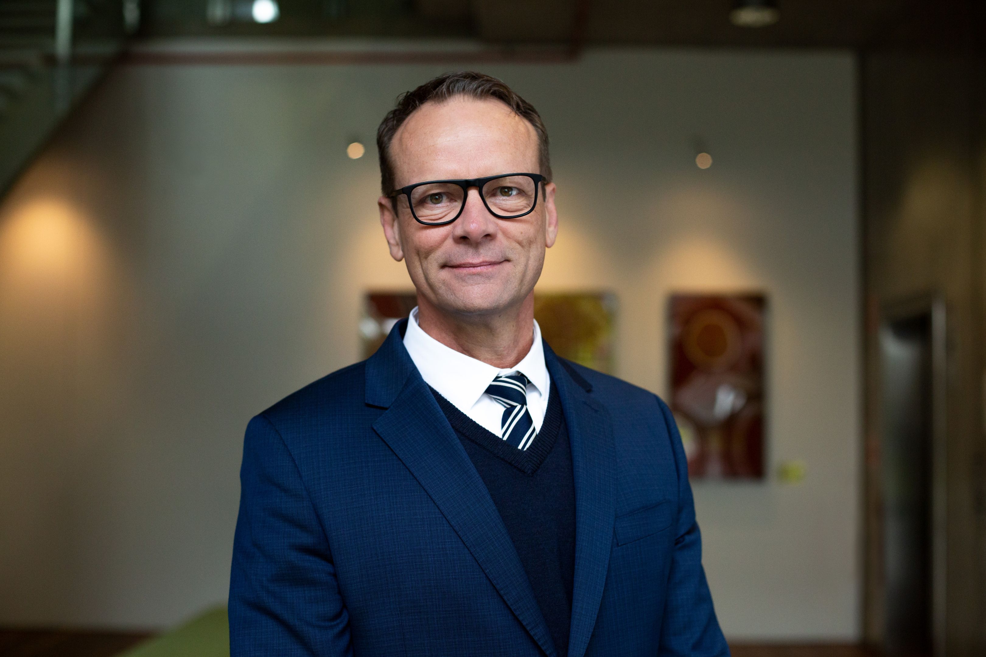 A male with short hair and glasses wearing a blue suit, white shirt and tie.