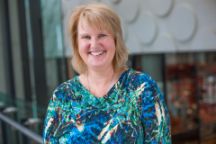 A blonde woman smiles to camera in front of a large building window wearing a blue blouse
