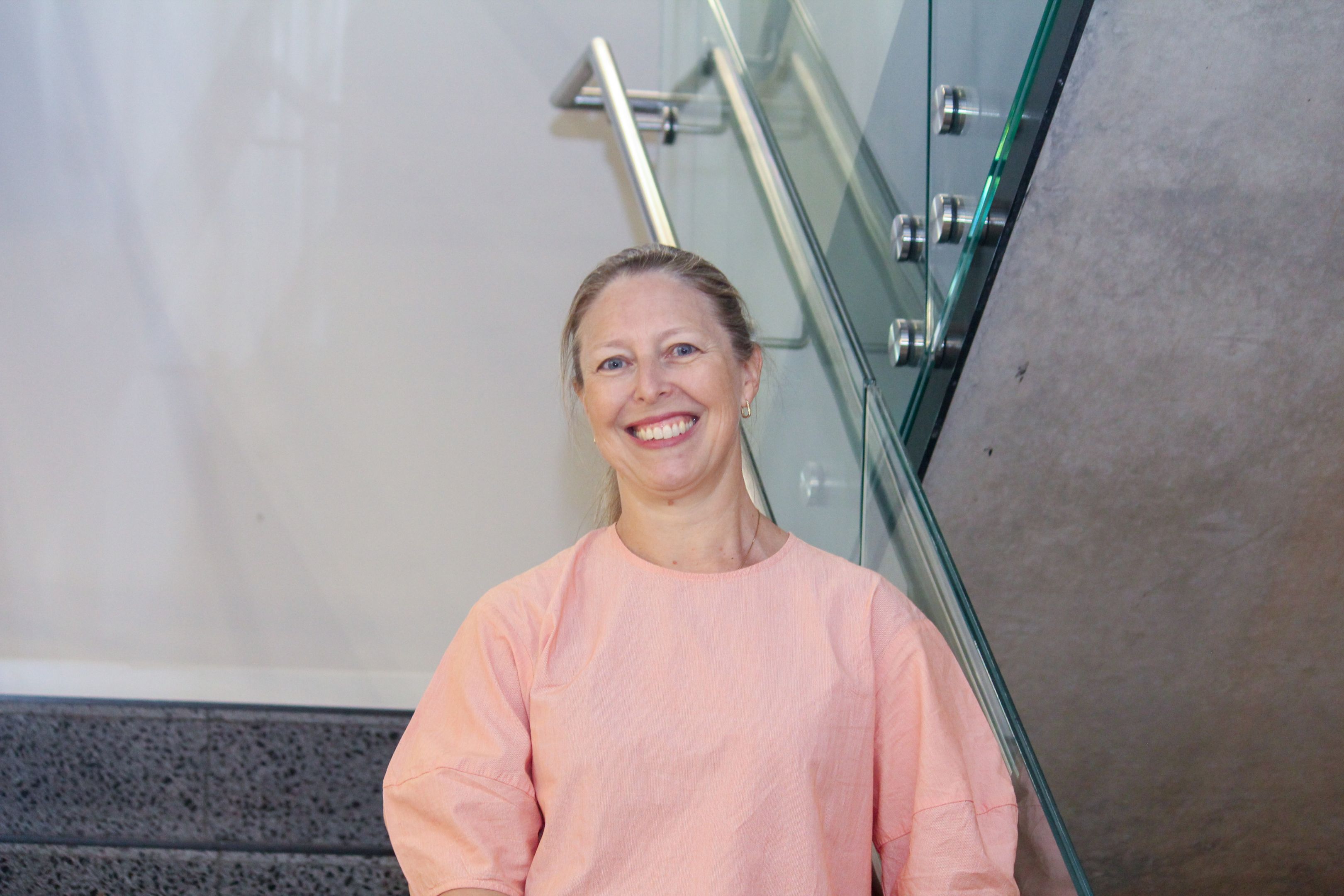 Swinburne Staff Member Tash Hobbs standing on a set of concrete stairs with a glass railing and a white wall behind. 