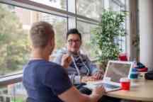 Two male students sit at a desk with a laptop and coffee cups