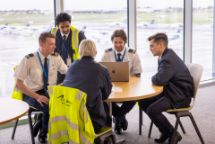 Aviation and piloting students sitting around a table with planes on the tarmac visible through the windows behind them.
