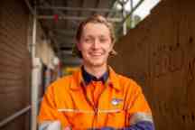 A smiling young man wearing an orange high-vis work shirt stands in front of a construction site