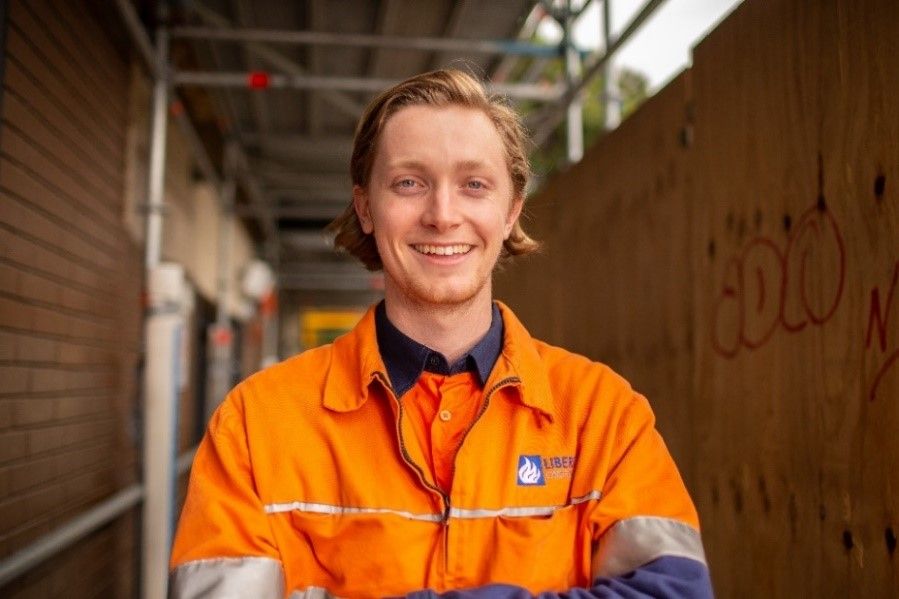 A smiling young man wearing an orange high-vis work shirt stands in front of a construction site